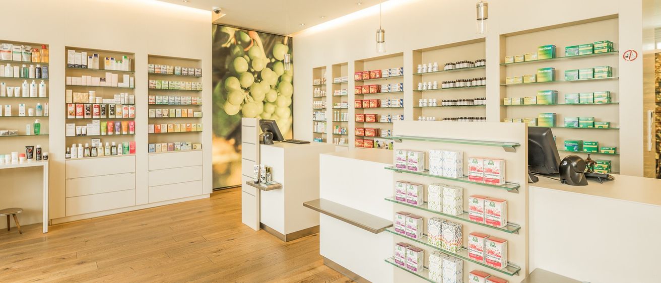 Eine Apotheke mit Holzböden und Regalen voller Produkte. Ein großes Poster mit Trauben ist an der Wand. Ein Schreibtisch hat einen Computermonitor und andere Gegenstände.