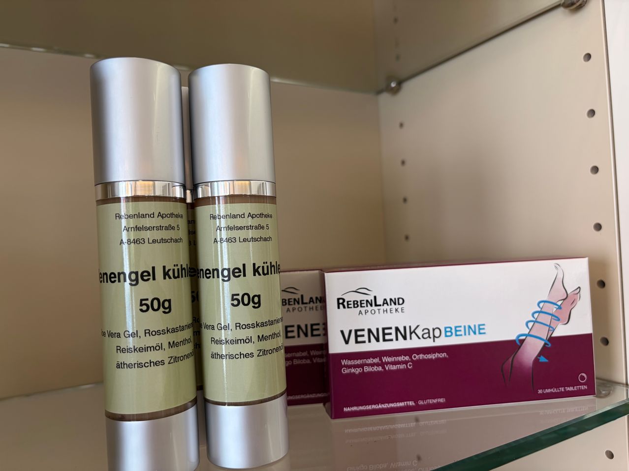 Two silver tubes labeled 'Rebenland Apotheke' are on a shelf with a box of 'VENENKap BEINE' nearby. The tubes contain 50g of gel, possibly with menthol and rosemary oil.