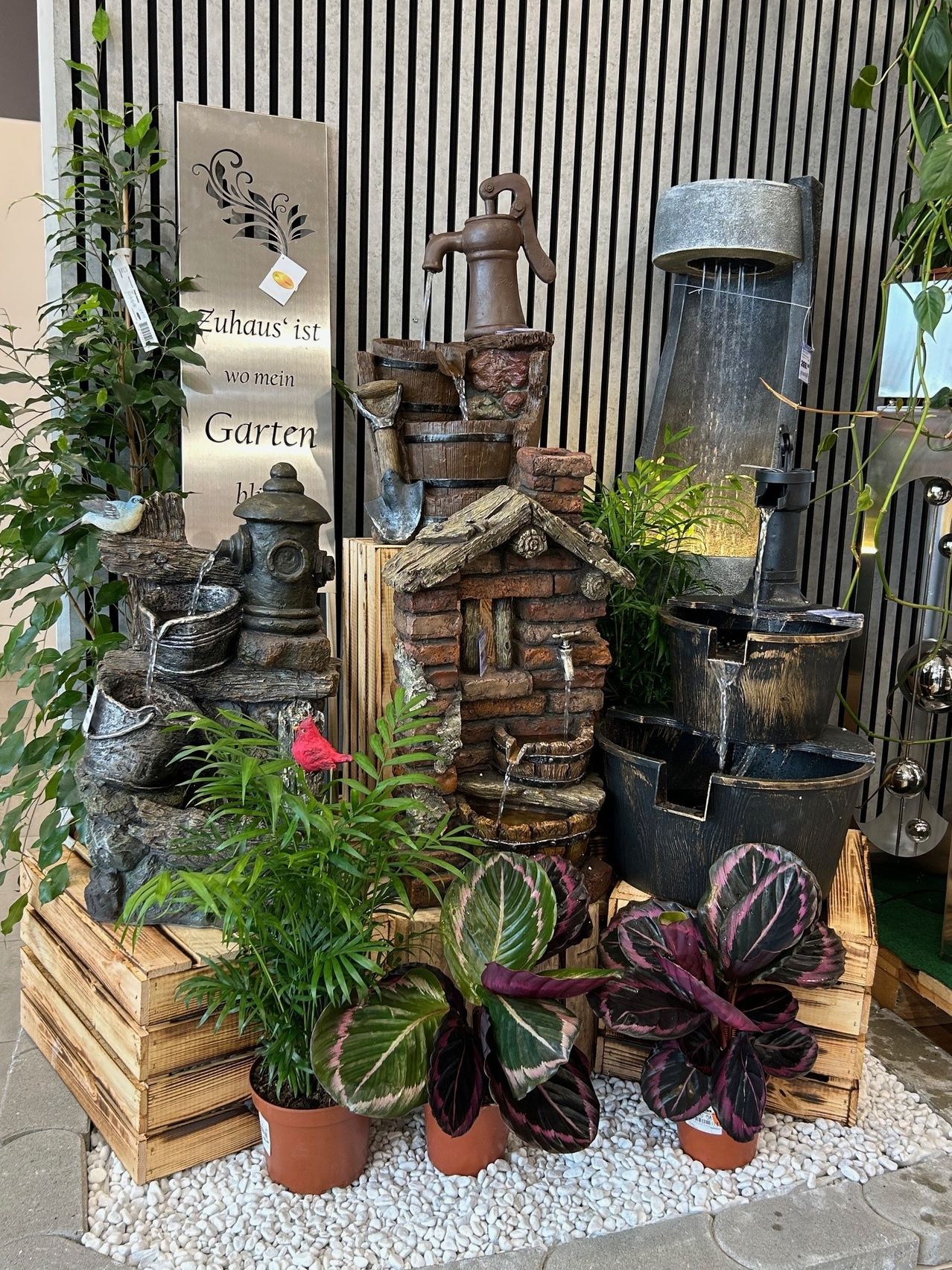 Eine Gartenausstellung mit einem Brunnen als Mittelpunkt, umgeben von verschiedenen Pflanzen und Dekorationsartikeln. Der Brunnen hat mehrere Ebenen mit fließendem Wasser.