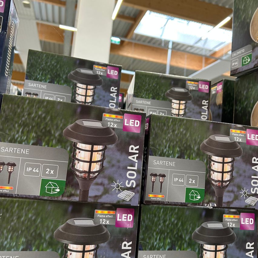 Mehrere Kartons mit LED-Solarleuchten sind in einem Geschäft ausgestellt. Die Kartons haben unterschiedliche Designs und Funktionen, einschließlich Flammeneffekt und IP44-Bewertungen. Die Produkte sind auf einem Tisch mit einem grünen Grashintergrund platziert.