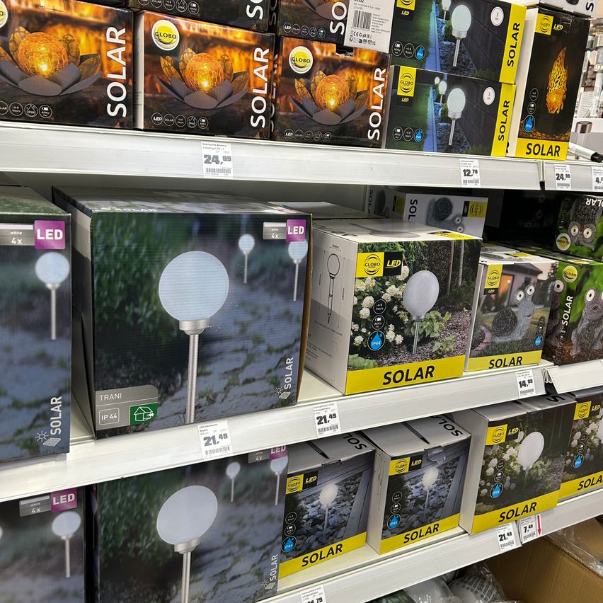 Ein Ladendisplay mit verschiedenen solarbetriebenen Gartenleuchten. Die Produkte sind ordentlich auf weißen Regalen angeordnet. Die Boxen zeigen Bilder von Solarleuchten und Preise sind sichtbar.