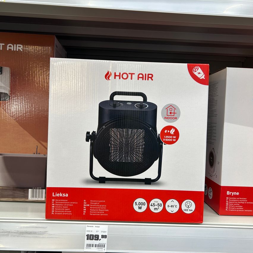 Eine weiße und rote Box für einen Heißluftheizer wird auf einem Regal ausgestellt. Die Box ist mit dem Markennamen 'HOT AIR' beschriftet und enthält ein Bild des Produkts. Die Box enthält Details über die Leistung und Sicherheitszertifikate des Produkts.