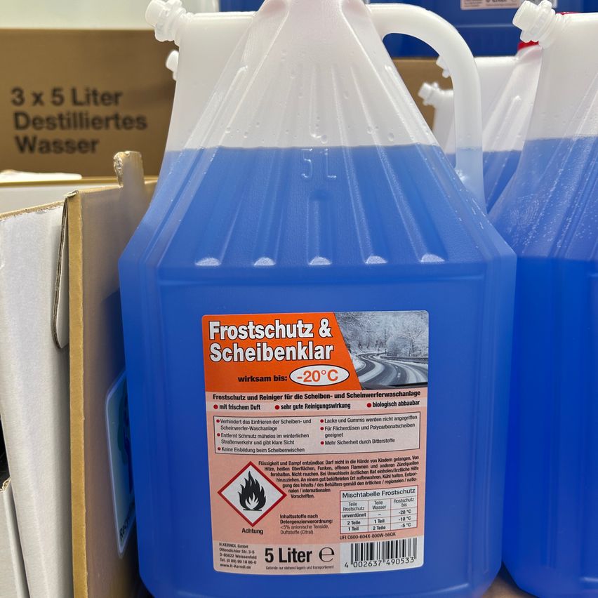 Eine blaue Plastikflasche mit einem Etikett, das 'Frostschutz & Scheibenklar' lautet, ist abgebildet. Sie ist mit einer blauen Flüssigkeit gefüllt, wahrscheinlich Frostschutzmittel. Das Etikett zeigt an, dass es bis -20°C vor Frost schützt und die Windschutzscheibe und das Sichtfenster reinigt.