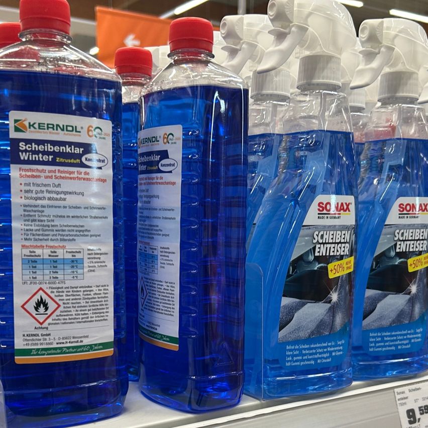 Mehrere Flaschen Scheibenwaschflüssigkeit stehen in einem Geschäft in einer Reihe. Sie sind blau mit roten Kappen und Etiketten mit Text in einer fremden Sprache. Die Flaschen stehen aufrecht und ordentlich angeordnet.