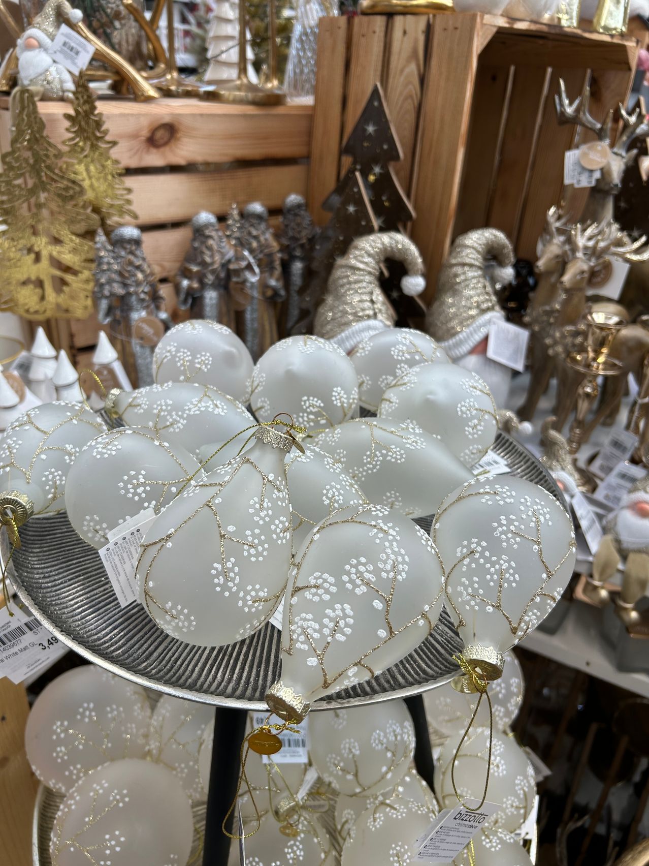 Ein Ladendisplay mit weißen Weihnachtskugeln mit goldenen Akzenten und filigranen Designs, auf einem mehrstufigen Ständer platziert, vor einem Hintergrund festlicher Dekorationen.