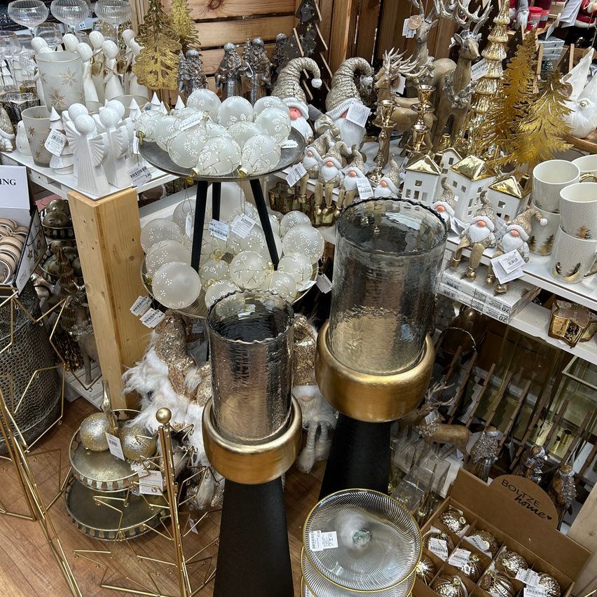 Eine Ausstellung von Dekorationsartikeln in einem Geschäft mit goldenen und silbernen Kerzenständern, weißen und goldenen Ornamenten und verschiedenen Glasartikeln auf Regalen. Im Vordergrund steht ein hölzerner Pfosten mit goldenen Akzenten.