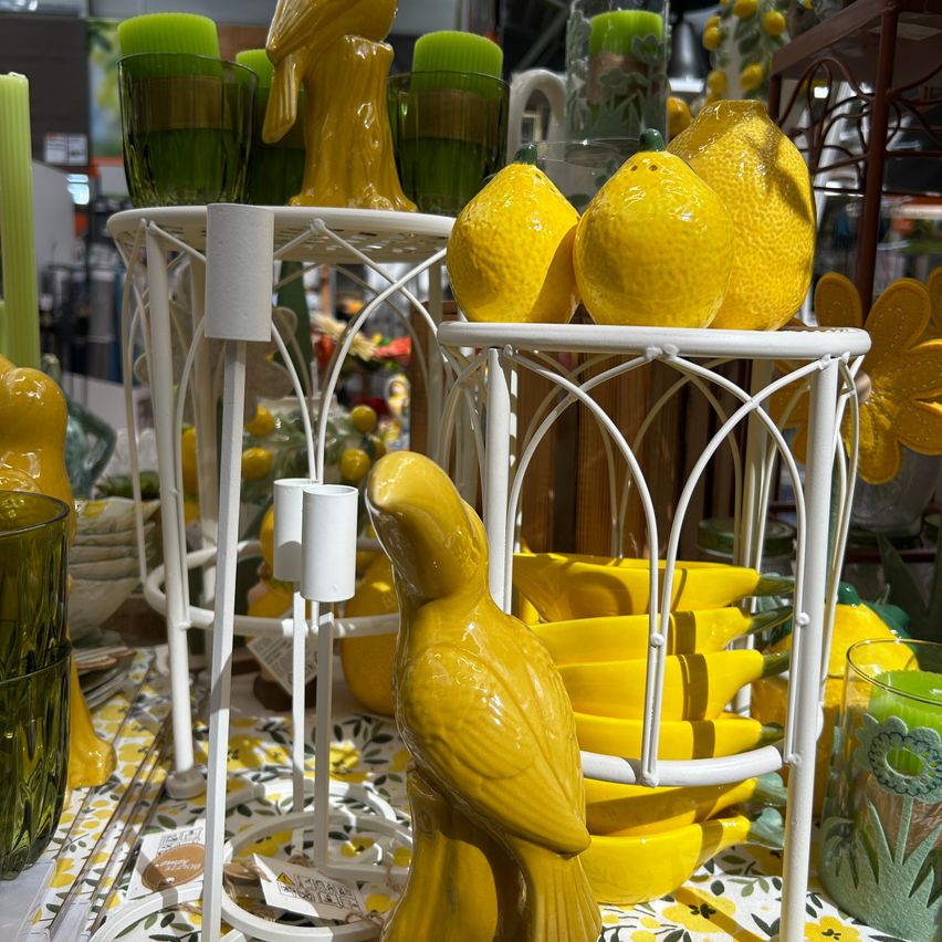 Eine Ausstellung von Dekorationsartikeln umfasst gelbe Keramikvögel, Zitronen und Kerzenständer auf weißen Ständern. Grüne Kerzen und Glasbecher sind ebenfalls vorhanden.