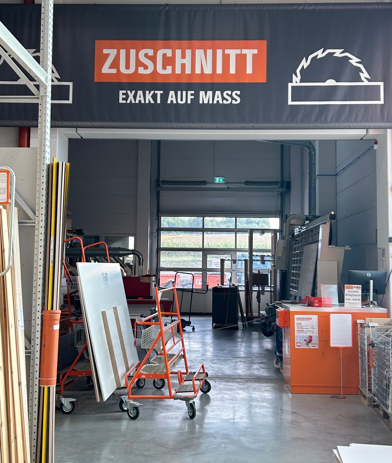 Der Eingang zu einem Geschäft zeigt ein Banner in orange und weiß. Das Banner sagt 'Zuschnitt' und 'EXAKT AUF MASS'. Der Laden hat Regale, Regale und Ausrüstung. Auf der rechten Seite befindet sich ein großes Fenster.