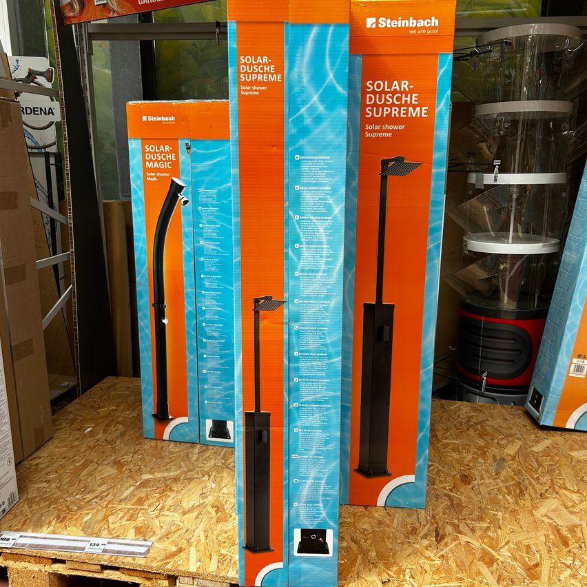 Drei Kartons mit Solar-Dusche Magic Solarduschen sind auf einer Holzoberfläche ausgestellt. Die Kartons sind orange und blau, mit dem Produktnamen und einem Bild eines schwarzen Duschkopfs.
