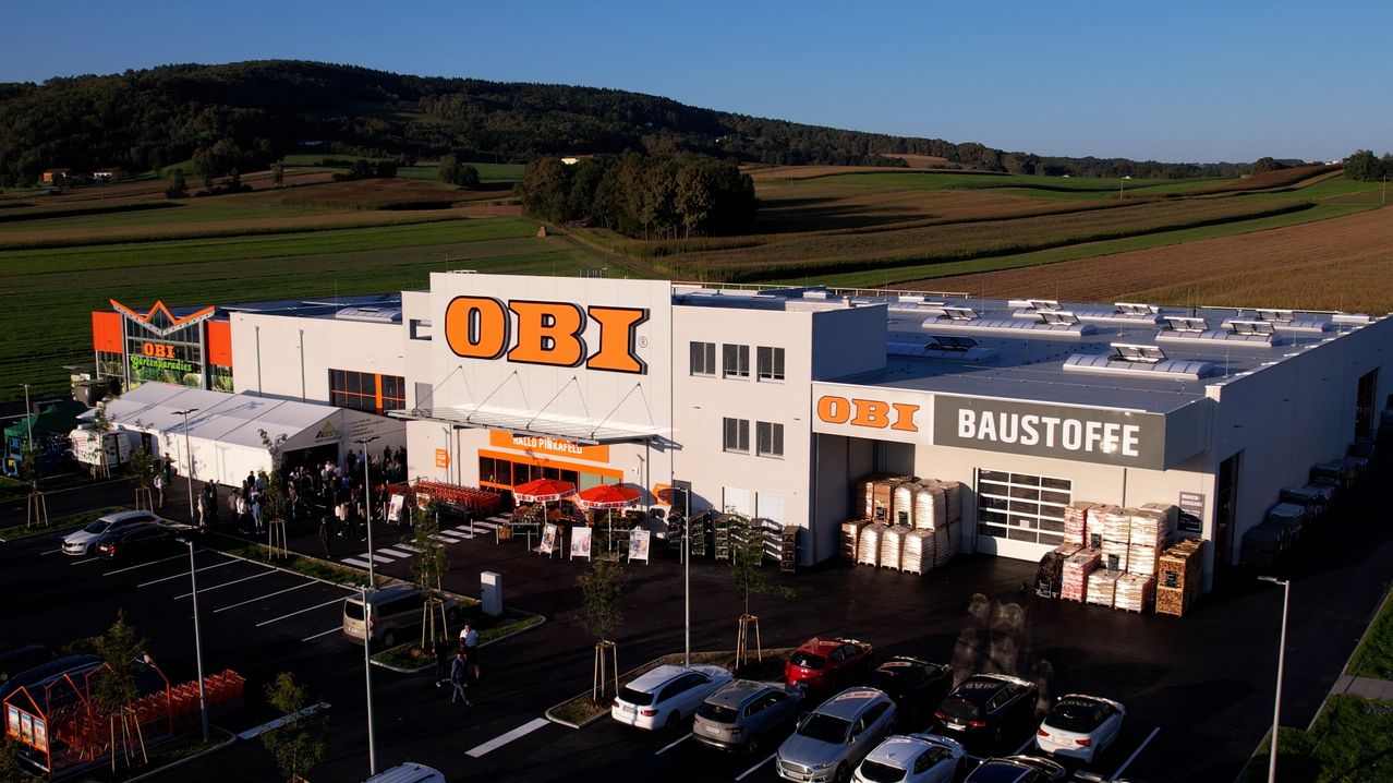 Eine Luftaufnahme eines OBI-Gebäudes mit mehreren Autos davor. Das Gebäude hat große orange Buchstaben und ist von einem Parkplatz umgeben.