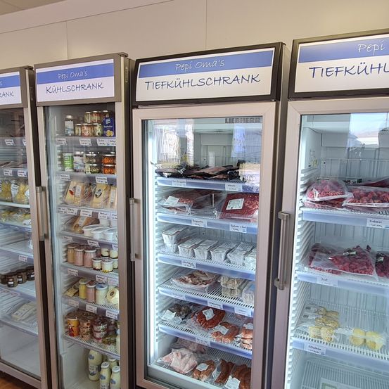 Drei große Kühlschränke sind an einer Wand aufgereiht. Jeder Kühlschrank ist mit verschiedenen Namen beschriftet. Die Kühlschränke enthalten verschiedene Artikel wie Fleisch, Käse und andere Lebensmittel.