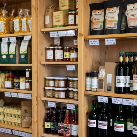 Ein hölzerne Regal präsentiert verschiedene Bio-Produkte, darunter Nudeln, Marmeladen, Öle und Gewürze. Eine Reihe von Weinflaschen und Gewürzen ist ebenfalls sichtbar.