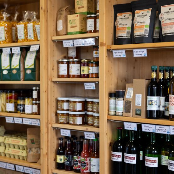 Ein hölzerne Regal präsentiert verschiedene Bio-Produkte, darunter Nudeln, Marmeladen, Öle und Gewürze. Eine Reihe von Weinflaschen und Gewürzen ist ebenfalls sichtbar.