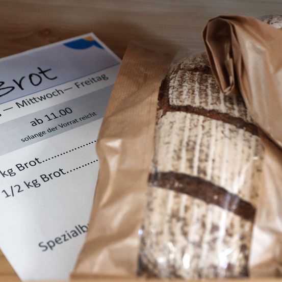 Ein braune Papiertüte mit Brot darin. Das Brot ist in Plastik eingewickelt. Ein Zettel sagt 'Brot - Mittwoch - Freitag, ab 11.00'. Gewichte von 1 kg und 1/2 kg sind aufgeführt.