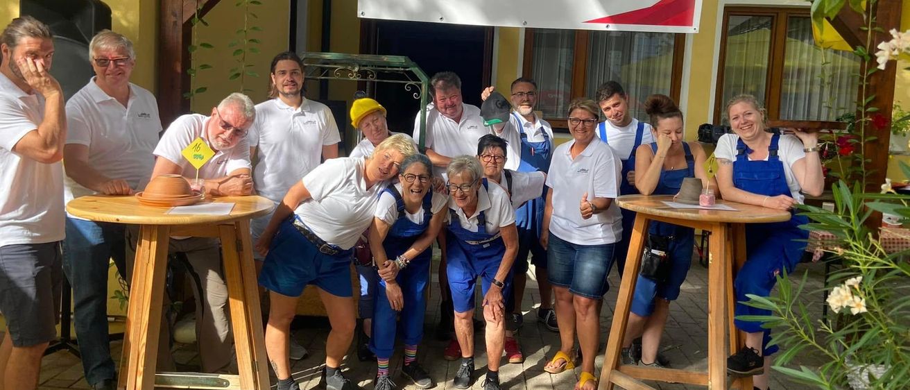 Eine Gruppe von Menschen in weißen Hemden und blauen Latzhosen posiert für ein Foto vor einem Gebäude mit einem Schild Herzlich Willkommen.