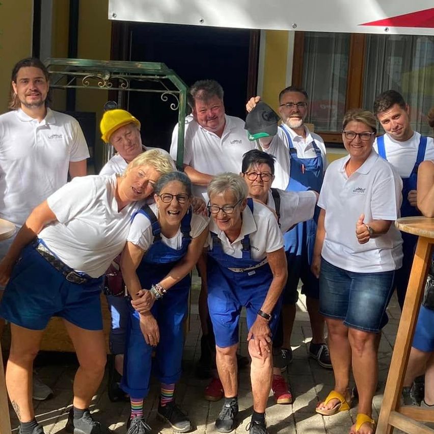 Eine Gruppe von Menschen in weißen Hemden und blauen Latzhosen posiert für ein Foto vor einem Gebäude mit einem Schild Herzlich Willkommen.