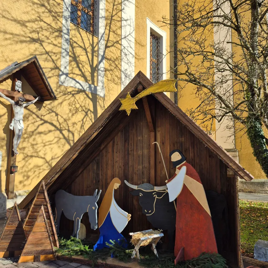 Eine Krippe vor einer Kirche mit Krippe, Figuren von Maria, Josef und Tieren sowie einem Stern darüber. Die Kirche hat gelbe Wände, Fenster und ein Kruzifix.