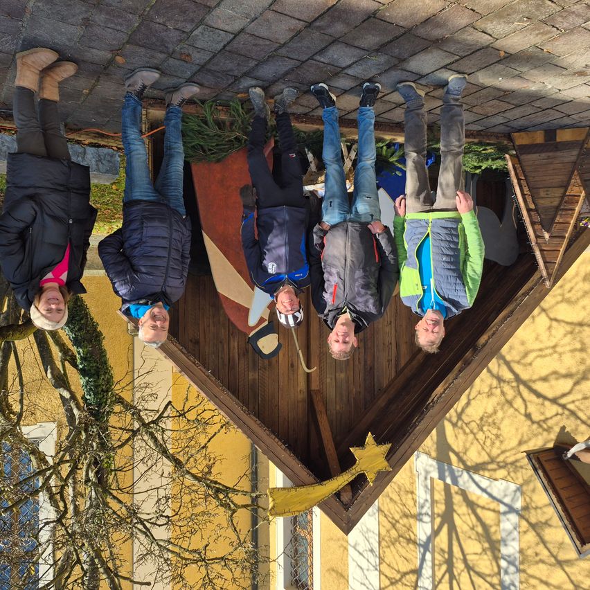 Fünf Personen stehen vor einer Holzstruktur, lächeln und posieren für ein Foto. Sie tragen Winterkleidung und Stiefel. Die Struktur hat einen gelben Stern am Boden.