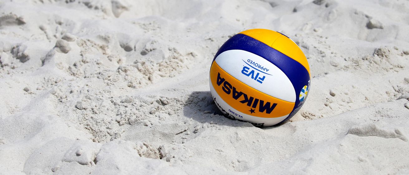 Ein Volleyball mit blauen, gelben und weißen Abschnitten liegt im Sand eines Strandes. Das Wort 'FIVB' ist in Blau geschrieben.