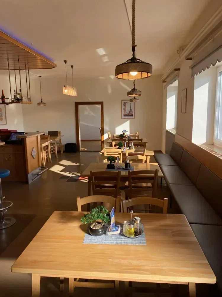 Ein leeres Restaurant mit Holztischen und Stühlen, Lampen und Pflanzen. Eine Bank steht in der Nähe der Fenster. Der Tresen hat Monitore, eine Flasche und andere Gegenstände.