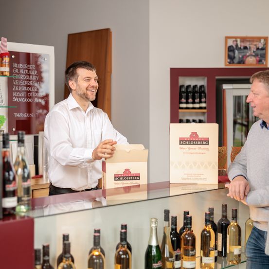 Ein Mann in einem weißen Hemd lächelt und steht hinter einer Theke in einem Weinladen. Er hält eine Schachtel Schlossberg-Wein und spricht mit einem anderen Mann. Der Laden hat eine Glastheke mit verschiedenen Weinflaschen und einem roten Schrank.