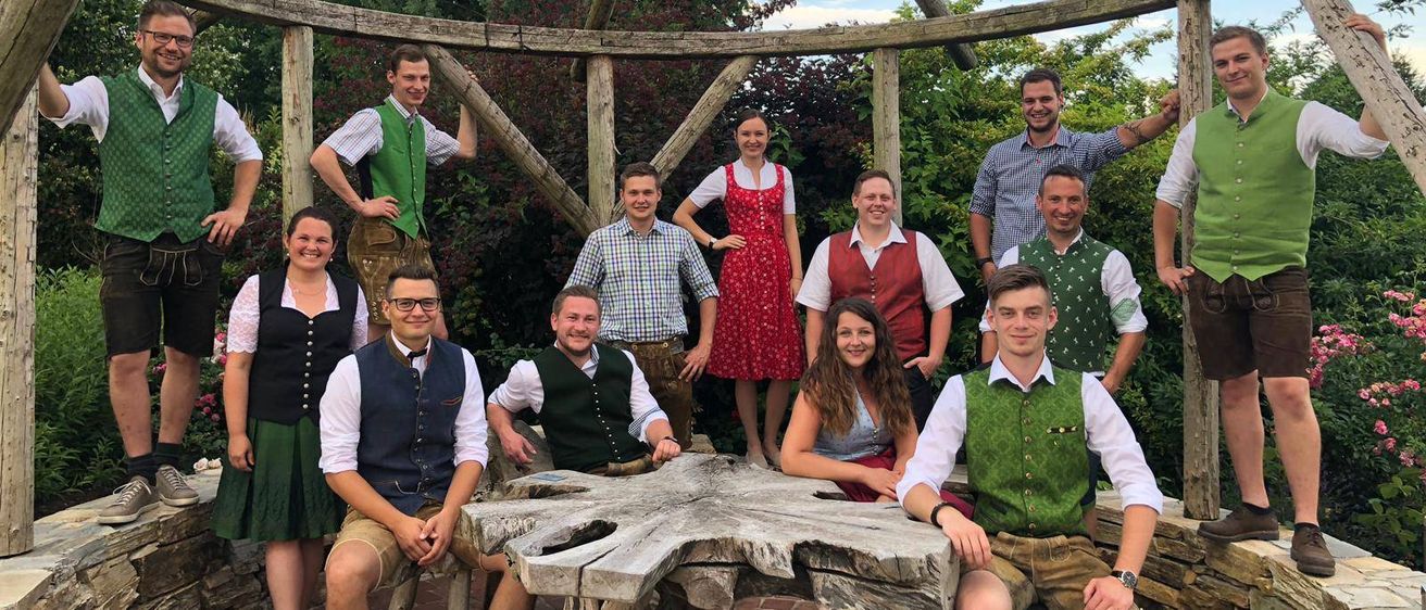 Eine Gruppe von Menschen in traditioneller Kleidung, die für ein Foto unter einem hölzernen Pavillon mit Ziegelboden posiert. Einige sitzen auf Bänken, während andere hinter einem Tisch stehen. Im Hintergrund sind Bäume zu sehen.
