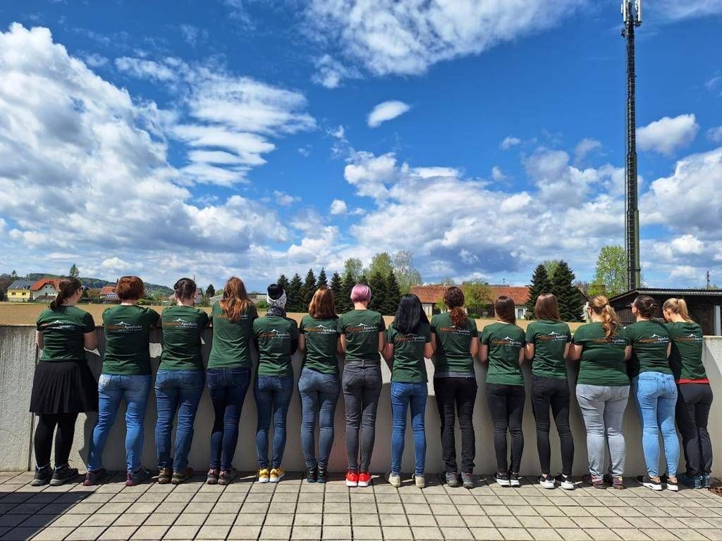 Eine Gruppe von Frauen in passenden grünen Shirts und Jeans steht in einer Reihe und schaut in die Ferne. Der Himmel ist blau mit verstreuten Wolken.