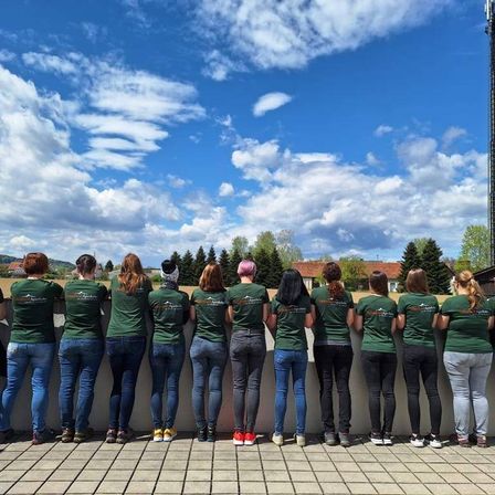 Eine Gruppe von Frauen in passenden grünen Shirts und Jeans steht in einer Reihe und schaut in die Ferne. Der Himmel ist blau mit verstreuten Wolken.