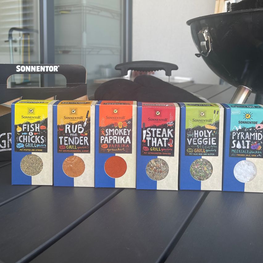 Sechs Schachteln Sonninator Gewürze stehen auf einem schwarzen Tisch. Die Schachteln enthalten 'Fish Chicks', 'Rub the Tender', 'Smokey Paprika', 'Steak That', 'Holy Veggie' und 'Pyramid Salt'.