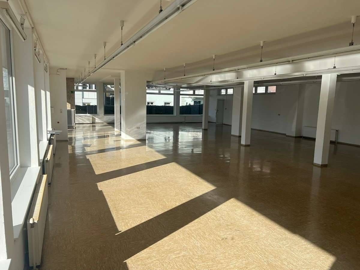 Ein leerer Raum mit Holzböden, weißen Wänden und hohen Fenstern. Sonnenlicht strömt herein und wirft Schatten auf den Boden. Der Raum ist sauber und unmöbliert.