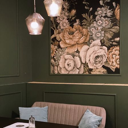Eine gemütliche Ecke mit floralem Wandpapier, einer gepolsterten Bank und zwei Lampen. Der Raum ist mit grünen Wänden und einer Holzbank geschmückt.