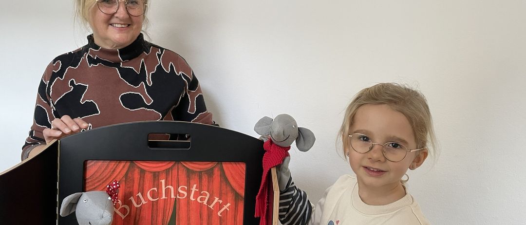 Eine Frau und ein Kind stehen neben einer schwarzen Box mit dem Wort Buchstart darauf. Die Frau lächelt und das Kind hält einen Kuscheltier.