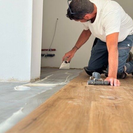 Ein Mann verlegt Holzboden auf einem Betonboden. Er benutzt eine Kelle, um Klebstoff aufzutragen.