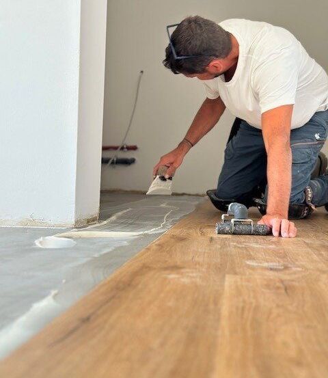 Ein Mann verlegt Holzboden auf einem Betonboden. Er benutzt eine Kelle, um Klebstoff aufzutragen.