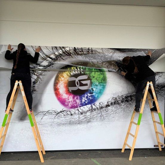 Zwei Frauen stehen auf Leitern und arbeiten an einer großen Wandmalerei eines Auges an einer Gebäudewand. Das Auge hat eine farbenfrohe Iris mit einer weißen Pupille.