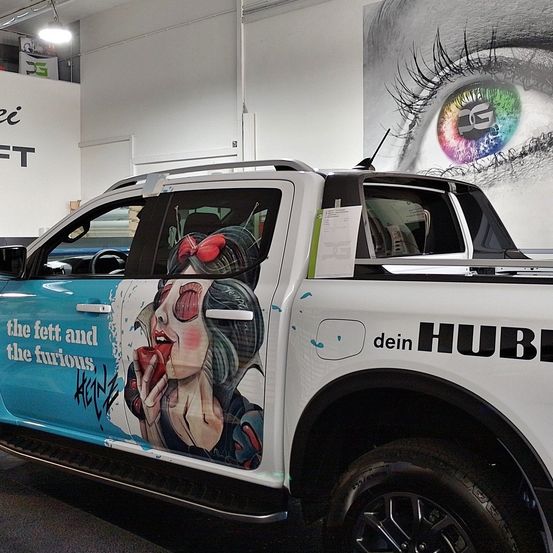 Ein weißer Pickup-Truck mit blauen Akzenten zeigt ein Bild einer Frau, die einen Apfel isst. Auf der Seite steht der Text 'the fett and the furious ACEN'. Er steht in einer Garage.