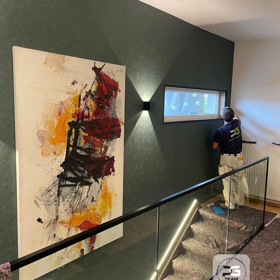 Ein Mann installiert ein großes abstraktes Gemälde an einer grünen Wand. Er steht auf einer Glastreppe neben einem Glasgeländer. Darüber befindet sich ein Fenster.