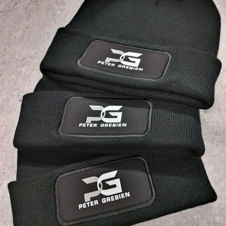 Drei schwarze Mützen gestapelt, jede mit einem weißen Logo, das 'DG PETER GREBIEN' auf einem schwarzen Aufnäher zeigt. Das Logo ist mit einem stilisierten 'DG' und 'PETER GREBIEN' darunter gestaltet.