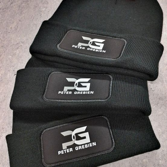 Drei schwarze Mützen gestapelt, jede mit einem weißen Logo, das 'DG PETER GREBIEN' auf einem schwarzen Aufnäher zeigt. Das Logo ist mit einem stilisierten 'DG' und 'PETER GREBIEN' darunter gestaltet.