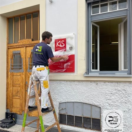 Ein Mann in einem blauen Shirt und weißen Hosen steht auf einer Leiter und bringt eine Plastikfolie auf einem roten Schild an, das an einer weißen Gebäudewand befestigt ist.
