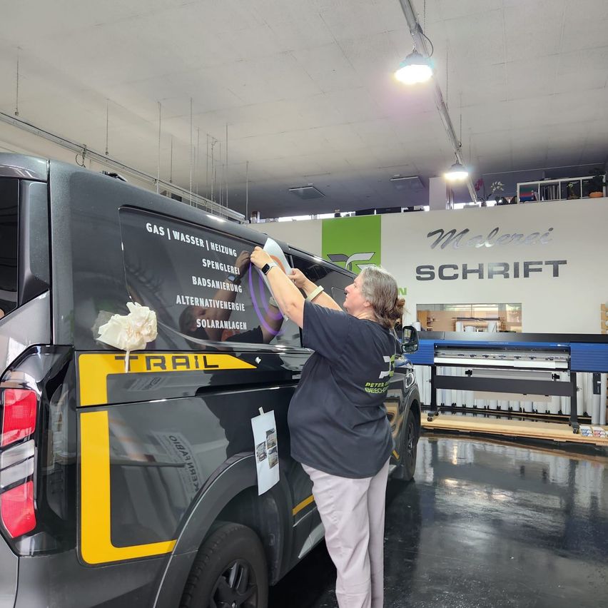 A woman is applying a sticker to the side of a black van with yellow trim. The van has text in German on its side.