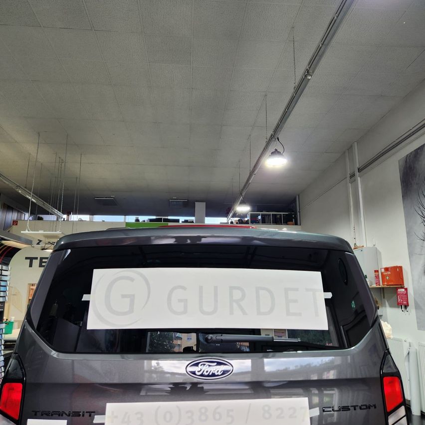 The back of a Ford Transit van with a sign reading 'G Gurdet' on its window. The van is in a garage with white ceiling and walls.