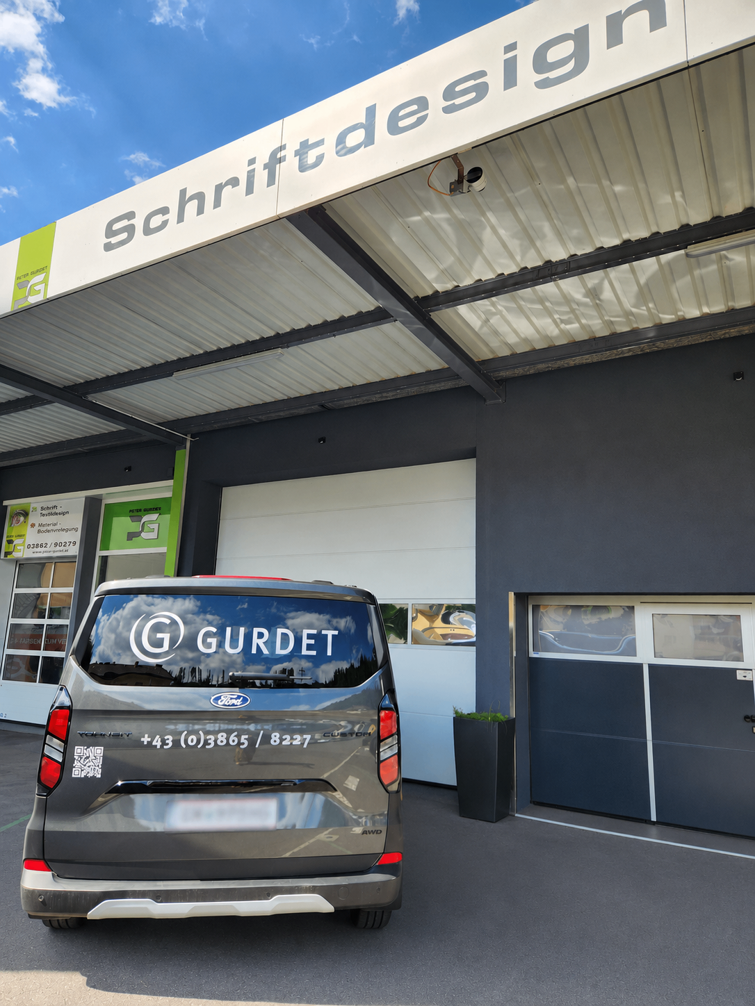 A Ford Transit van with the Gurdet logo on the back is parked in front of a building with a gray wall and white roof. The building has a sign that reads 'Schriftda' and a surveillance camera on the ceiling.