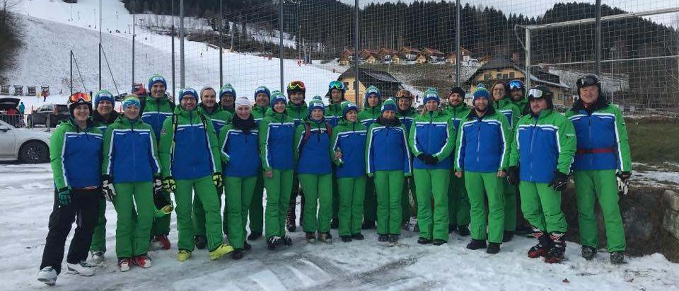Eine Gruppe von Menschen in passender grüner und blauer Winterkleidung posiert für ein Foto im Schnee. Dahinter befindet sich ein Zaun mit einem Torpfosten. In der Ferne sind Häuser und ein verschneiter Berg zu sehen.