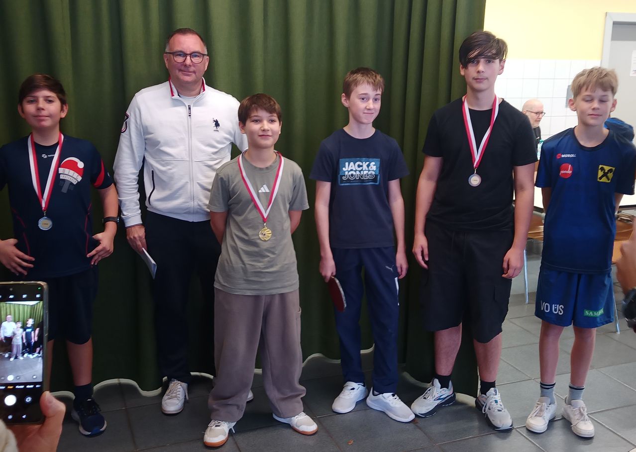 Fünf Jungen stehen gemeinsam vor einem grünen Vorhang und tragen Medaillen. Drei von ihnen tragen schwarze T-Shirts, einer trägt eine weiße Jacke und einer trägt ein graues Hemd. Sie alle lächeln.