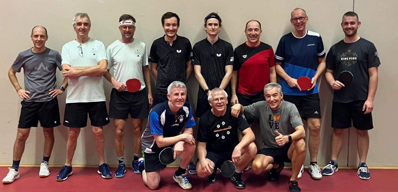 A group of people are posing for a photo. They are holding ping pong paddles. The people are wearing different shirts, shorts, and shoes. They are smiling and seem to be posing for a group photo.