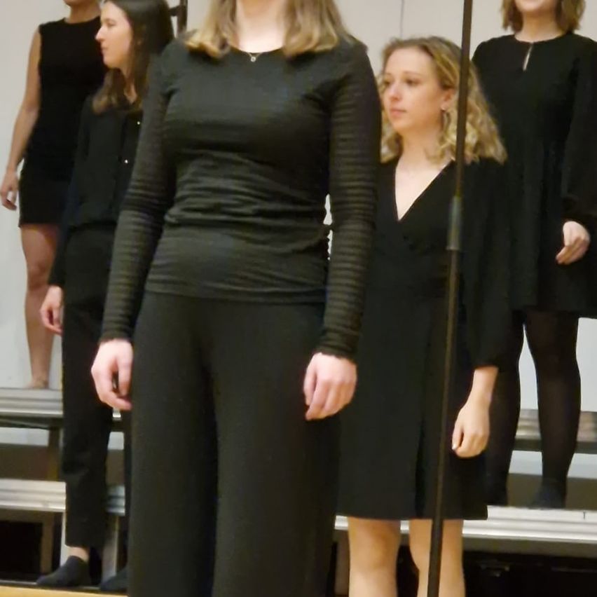 Fünf Frauen in Schwarz stehen auf einer Bühne, wahrscheinlich in Vorbereitung auf eine Aufführung. Die Frau in der Mitte trägt eine Halskette und weite Hosen.