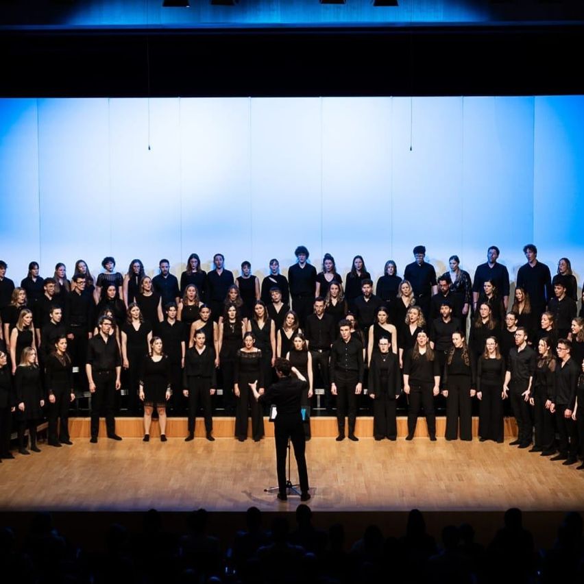 Ein Chor in schwarzer Kleidung tritt auf einer Bühne mit blauem Hintergrund auf. Ein Dirigent steht vor ihnen und gestikuliert. Das Publikum sitzt vor der Bühne.