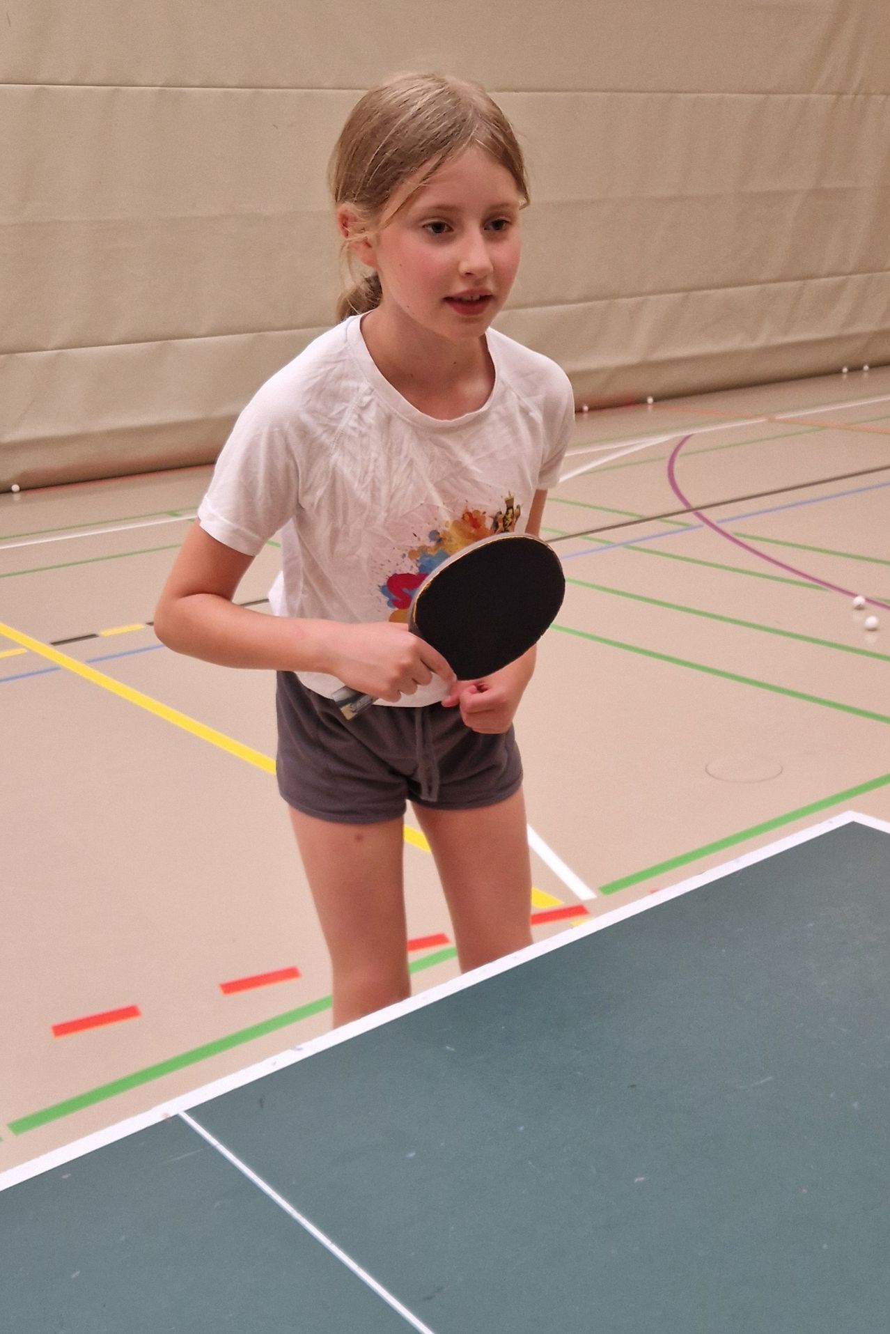 Ein junges Mädchen hält eine Tischtennisschläger in einer Turnhalle, bereit zum Spielen.