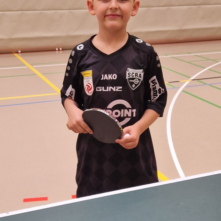Ein junger Junge steht in einer Sporthalle, hält eine Tischtennisschläger und einen Ball und trägt ein schwarzes Sporttrikot mit verschiedenen Sponsorenlogos.